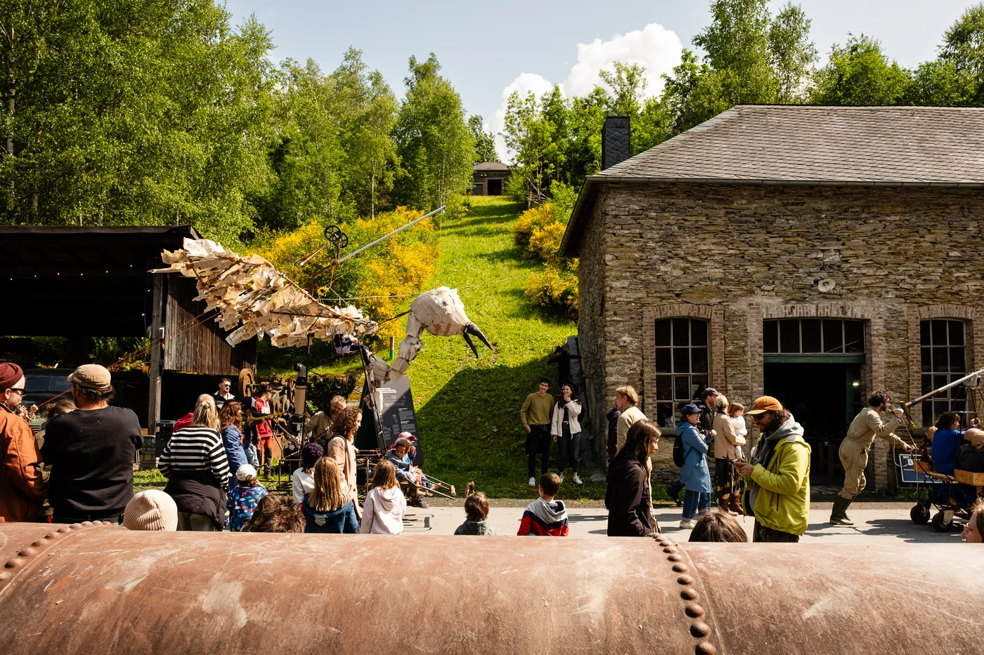 Festivalstemning ved steinbygningene på Koll an Aktioun, med publikum og en stor figur i oppvisningen.
